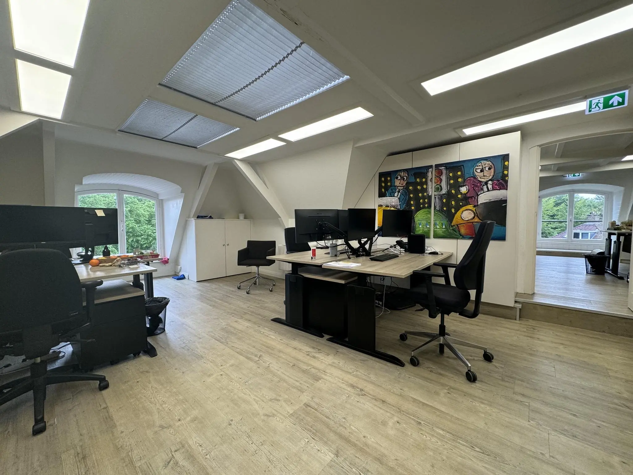 Modern kantoor op zolderverdieping met bureaus, computers en kleurrijk schilderij aan de muur.